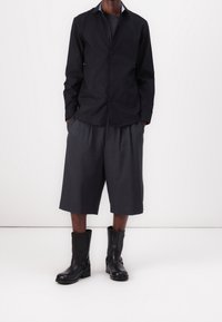 Camicia nera con bottoni e collo, abbinata a pantaloncini grigi plissettati e stivaletti neri in pelle con fibbie.