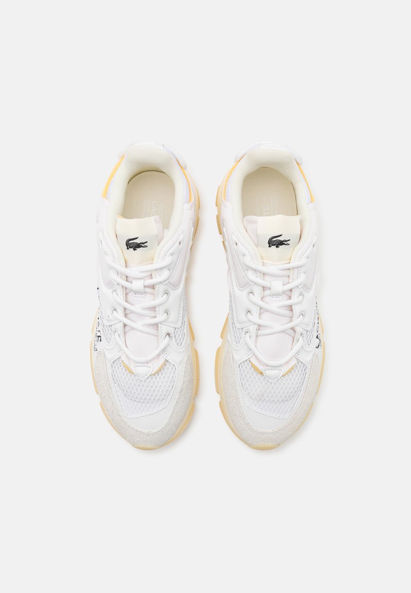 Zapatillas deportivas blancas con paneles de malla y gamuza, cuellos acolchados y suelas de goma amarilla; presenta un logotipo negro en la lengüeta.