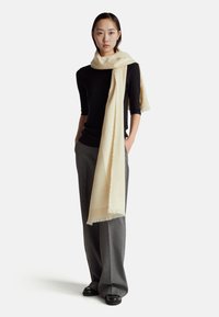 Béžový šátek z jemné, lehké látky, volně přehozený přes krk. Modelka má na sobě černý top a šedé kalhoty, s čistým, moderním vzhledem.