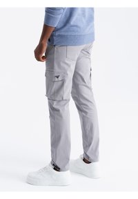 Grijze cargobroek met meerdere grote zakken en subtiele vouwdetails. Gepaard met witte sneakers en een lichtblauw overhemd met lange mouwen.