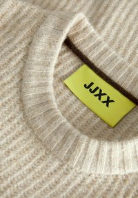 Suéter beige con textura, cuello redondo con canalé y una etiqueta amarilla con la inscripción "JXXX" adherida al interior del cuello.