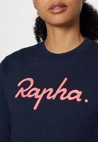 Gros plan d'une personne portant un sweat-shirt bleu marine à col rond avec un texte rose en écriture cursive "Rapha" sur la poitrine.