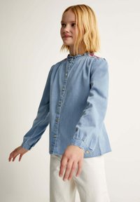 Cămașă din denim de culoare albastră deschisă, cu guler fronsat, închidere cu nasturi în față și broderie florală pe umeri. Mâneci lungi cu manșete cu nasturi.