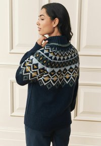 Tumsāli zils adīts džemperis ar ribainu apkakli un krāsainām ģeometriskām joslām zaļā, dzeltenā un pelēkā krāsā uz pleciem.
