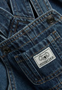 Denim tuinbroeken met blauwe stof, oranje stiksels en een logopatch met een koala-illustratie. Bevat een voorzak en metalen hardware.