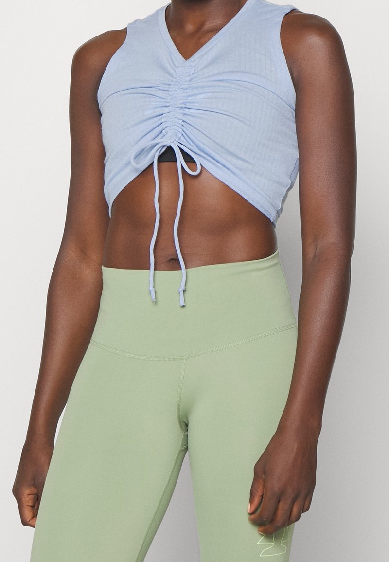 Buste féminin portant un crop top froncé bleu clair avec cordons et des leggings vert clair taille haute, debout devant un fond uni.