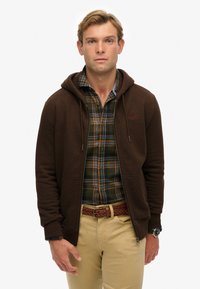 Sudadera marrón con cremallera y capucha ajustable, llevada sobre una camisa de cuadros verde y azul, combinada con pantalones color beige y un cinturón trenzado.