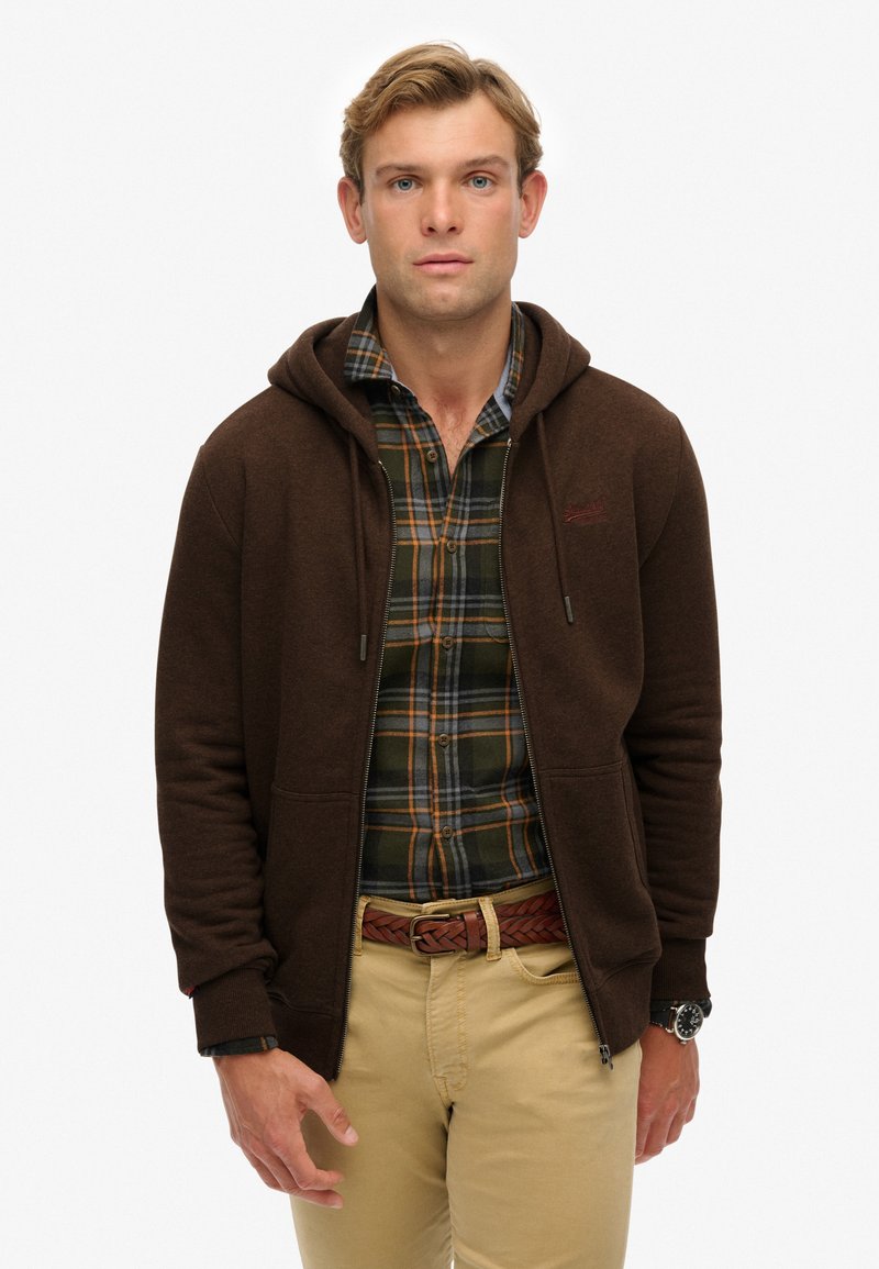 Sudadera marrón con cremallera y capucha ajustable, llevada sobre una camisa de cuadros verde y azul, combinada con pantalones color beige y un cinturón trenzado.