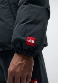 Black jacket with elastic cuff, featuring a red square logo patch labeled "The North Face" on the sleeve. Smooth, lightweight fabric.