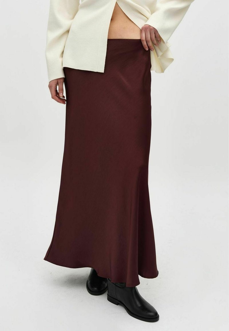 Lunga gonna di satin bordeaux con silhouette fluida, caratterizzata da una vita semplice e una texture liscia, abbinata a stivaletti neri alla caviglia.