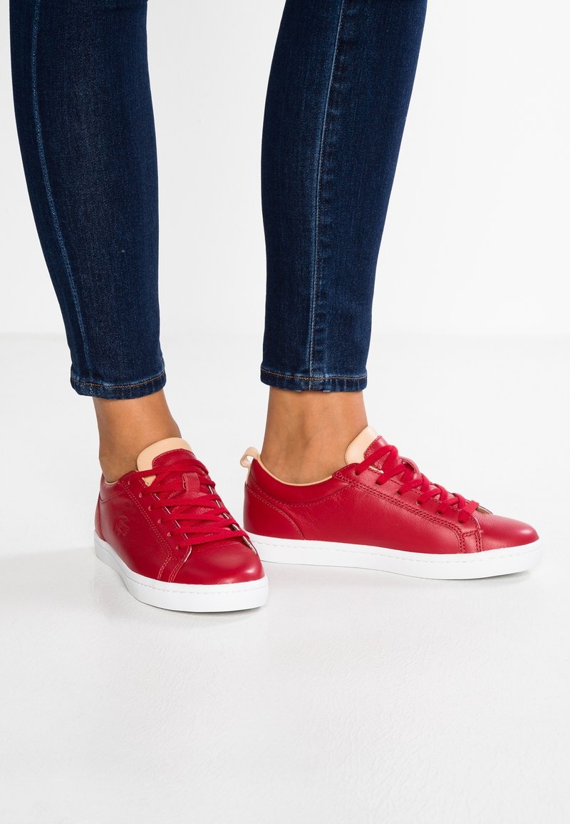 Fötter i röda lädersneakers med beige tunga och vita sulor, kombinerade med mörkblå skinny jeans mot en vit bakgrund.