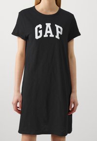 Černé bavlněné šaty ve tvaru trička s krátkými rukávy, kulatým výstřihem a bílým logem "GAP" napsaným tučným písmem napříč hrudí.