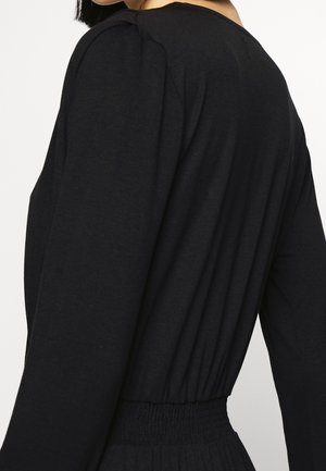 Woman wearing a black long-sleeve top with gathered elastic waist and subtle shoulder pleats, shown from the back and side.