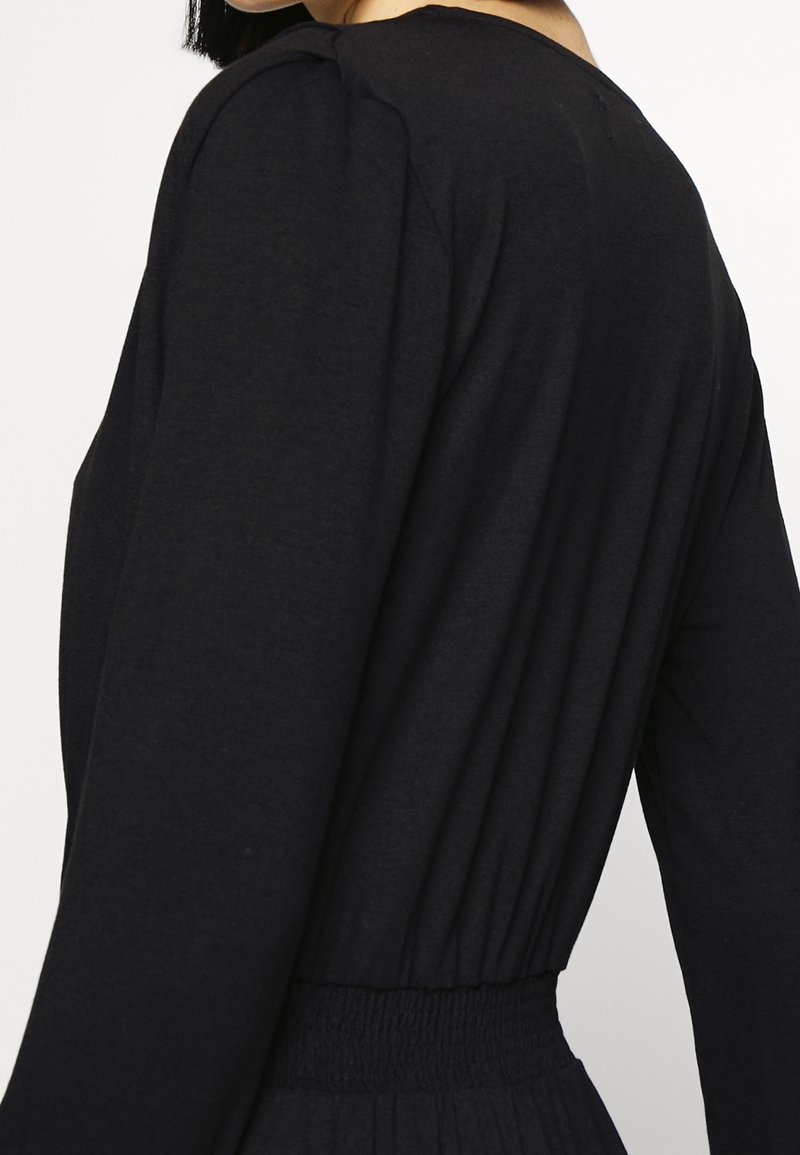 Femme portant un haut noir à manches longues avec une taille élastique froncée et de subtils plis aux épaules, vue de dos et de profil.