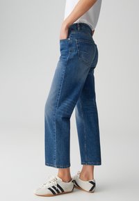 Jean en denim bleu avec une coupe décontractée et droite, présentant une teinte moyenne, cinq poches et un effet délavé subtil. Porté avec des chaussures blanches.