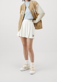 Lichtbruine varsityjas met witte mouwen, een hemelsblauw cropped shirt, een witte geplooide rok en witte sneakers met beige accenten.