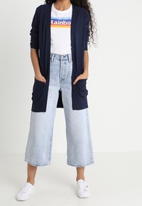 Marinblå cardigan med två stora framfickor, lager-på-lager över en vit t-shirt med en regnbågsdesign, i kombination med ljusblå vida jeans och vita sneakers.