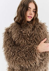 Mladá žena s hnědými vlasy po ramena, oblečená v tlustém, kudrnatém hnědém kožichu, se zkříženýma rukama, neutrální pozadí.