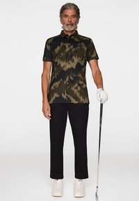 Pikétröja med kamouflagemönster i mörkgrönt och svart, kortärmad, matchad med svarta byxor och vita skor, håller i en golfklubba.