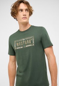 Young man avec des cheveux bouclés portant un T-shirt vert arborant un logo vintage "MUSTANG" imprimé en beige et blanc sur la poitrine.