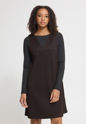 Jeune femme aux cheveux bouclés porte une robe sans manches marron foncé par-dessus un haut noir à manches longues, debout devant un fond clair uni.