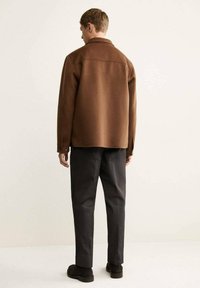 Giacca camicia in mélange di lana marrone con taglio dritto, polsini con bottoni e design con yoke posteriori. Abbinata a pantaloni scuri e scarpe marroni.