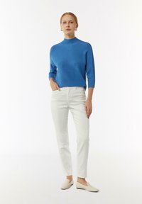 Maglione blu a maglia con collo alto, maniche 3/4, pantaloni bianchi aderenti e scarpe slip-on beige chiaro, su uno sfondo bianco semplice.
