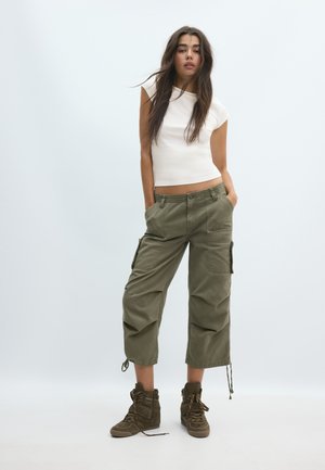 Jeune femme aux longs cheveux bruns portant un haut blanc sans manches, un pantalon cargo vert olive et des baskets montantes marron, debout les mains dans les poches.