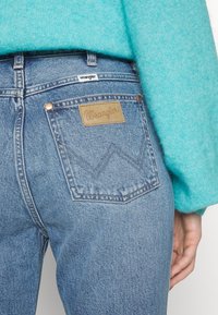Jean en denim bleu taille haute avec une étiquette en cuir beige portant la mention "Wrangler". Les poches arrière présentent un motif chevron brodé.