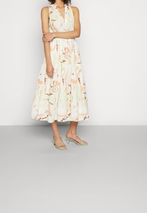 Frau steht mit verschränkten Armen, trägt ein ärmelloses Midikleid mit Blumenmuster und hellgrüne, gepolsterte Sandalen mit Absatz, vor einer schlichten Wand.