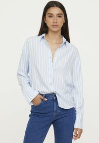 Light blue and white striped long-sleeve button-up shirt with a collar and single button closure. Paired with blue jeans.