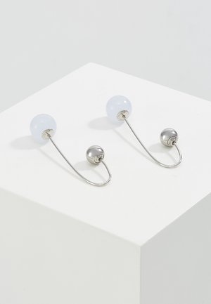 Par af sølv- og hvide dobbeltsidede ørestikker med en buet metalstang, der forbinder de to kugler, på en hvid overflade.