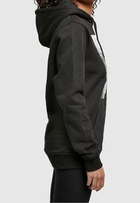 Osoba stojící bokem, oblečená v černé mikině s velkým bílým grafickým motivem na hrudi a černých legínách, na neutrálním pozadí.