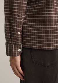 Ruda languota marškiniai su baltais mygtukais rankogaliuose, pagaminti iš tekstūruotos medžiagos. Derinami su tamsiai rudomis kelnėmis, kurios turi užpakalinę kišenę.