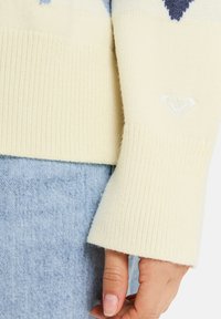 Kremfarget strikket genser med ribbestrikkede mansjetter, med et subtilt hjerteemblem, kombinert med lyseblå denim. Myk tekstur og geometrisk design.