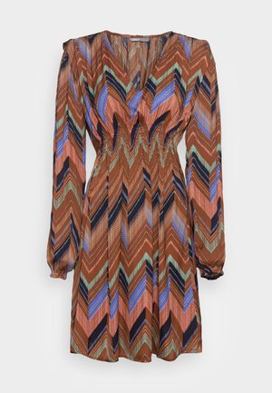 Brązowa sukienka o długości do kolan z długim rękawem, dekoltem w serek oraz wielokolorowym, zygzakowatym wzorem w odcieniach niebieskiego, zielonego, czarnego i różowego.