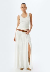 Witte ribbelige tanktop gecombineerd met een vloeiende witte maxi-rok met een zijsplit, een bruine riem en beige enkellaarsjes.