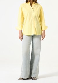 Żółta koszula na guziki z długimi rękawami i kieszenią na piersi, zestawiona z jasnoniebieskimi dżinsami o szerokich nogawkach oraz metalicznymi butami.