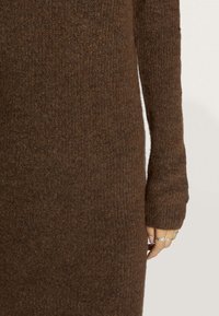 Brown knitted sweater with a textured, ribbed design; features long sleeves and a fitted silhouette. Hand shows silver rings.