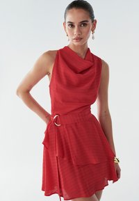 Punainen mekko, jossa on kaulus, hihaton malli, kerroksellinen helmassa ja kultainen renkaallinen solki vyötäröllä. Valmistettu teksturoidusta kankaasta.