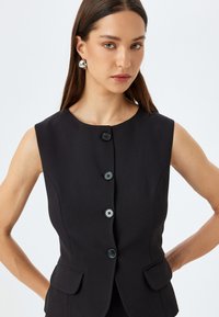 Zwarte mouwloze vest met een ronde halslijn, vijf zwarte knopen en zijzakken. De stof heeft een gladde textuur en een getailleerd ontwerp.