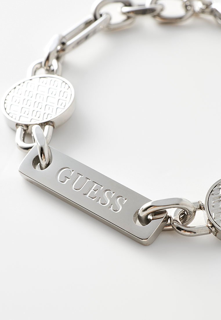 Bracelet en chaîne en métal argenté avec des maillons alternés, plaque gravée avec le logo "GUESS" et accents ronds avec un motif texturé.