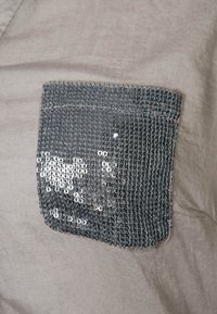 Tessuto grigio con una tasca testurizzata decorata con paillettes argento. La tasca presenta un bordo e è cucita sul capo.