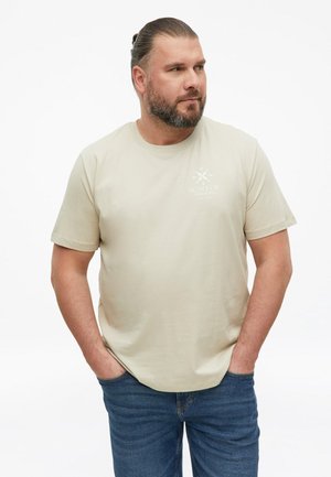 Man with beard and tied-back hair wearing a beige T-shirt with a small logo and blue jeans, standing with hands in pockets.
