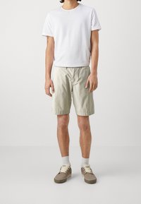 T-shirt bianco a maniche corte, leggermente testurizzato. Pantaloni corti beige chiaro con elastico in vita. Scarpe marroni con lacci crema e calzini grigi.