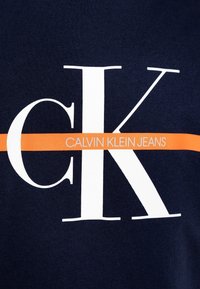Námornícky modré bavlněné tričko s velkým bílým logem "CK" a oranžovým pruhem s textem "CALVIN KLEIN JEANS" pod ním.