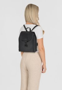 Zwarte leren rugzak met een klep-sluiting, trekkoorddetail en verstelbare banden, getoond op een persoon die een wit T-shirt en beige broek draagt.