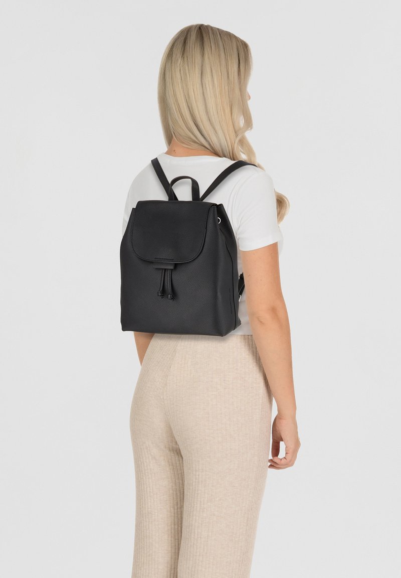 Zwarte leren rugzak met een klep-sluiting, trekkoorddetail en verstelbare banden, getoond op een persoon die een wit T-shirt en beige broek draagt.