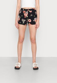 Femme portant un short noir à motifs floraux et un haut blanc, debout pieds nus sur un sol gris clair contre un mur blanc.