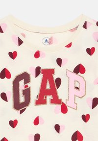Μπλούζα για νήπια σε κρεμ χρώμα με κόκκινες, ροζ και μαρόν καρδιές και γράμματα με λάμψη που σχηματίζουν το GAP στο στήθος.